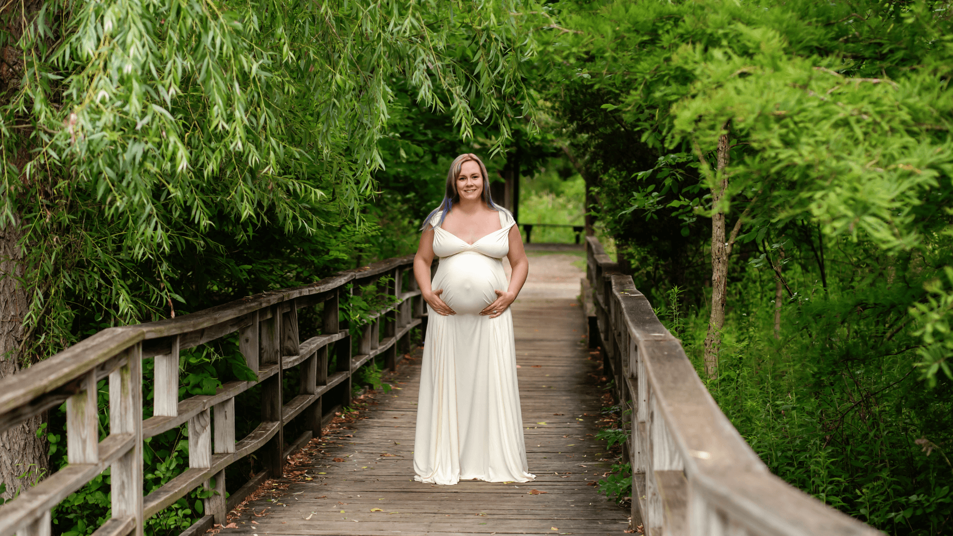 Pregnant woman wearing a white dress and standing on a wooden bridge in Ann Arbor, Michigan.